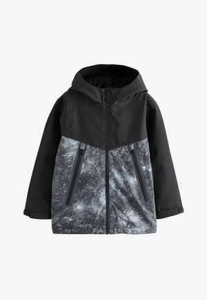 Młoda osoba stoi, nosi czarno-szarą marmurkową bluzę na zamek z kapturem, jasnoszare spodnie dresowe i białe buty sportowe z brązowym znakiem.