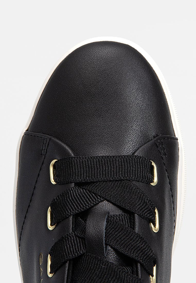 Svart lädersneaker med rund tå, utrustad med svarta elastiska snören och ögla i guldton, placerad mot en vit gummisul.