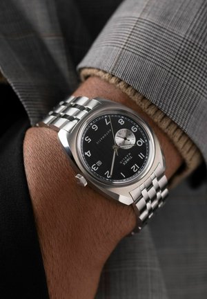 Montre automatique argentée avec un cadran noir, grands index horaires blancs, fenêtre de date et bracelet en métal. Portée sur un poignet avec une manche texturée.