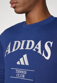 Blå sweatshirt med rund hals. Har hvid broderet tekst "ADIDAS" og "TENNIS CLUB" med logoet med tre striber nedenfor. Blød tekstur.