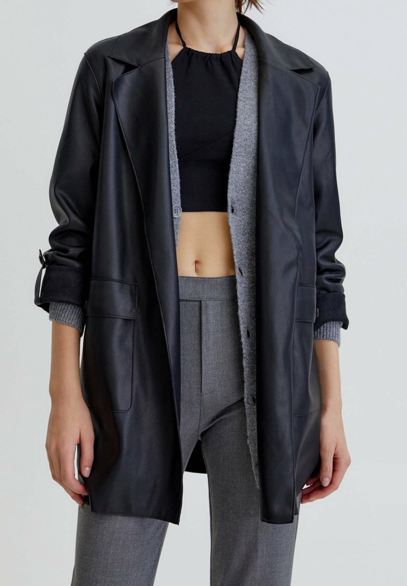 Personne portant une tenue superposée composée d'un haut court noir, d'un cardigan gris, d'une veste en cuir noire et d'un pantalon gris sur mesure, debout devant un fond uni.