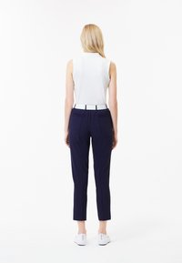 Witte mouwloze top, marineblauwe broeken met een aansluitende pasvorm, witte riem als accent, gladde textuur en witte instapschoenen, gezien vanaf de achterkant.