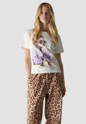 Blonde Frau mit langen Haaren trägt ein weißes T-Shirt mit einer Grafik einer Frau mit Sonnenbrille und beige Leopardenmuster-Hosen mit Taschen.