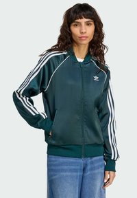 Grön satinfärgad sportjacka med vita ränder längs ärmarna, dragkedja framtill och sidofickor. Ribbstickade muddar och fåll, logotyp på bröstet.