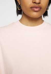 Sweatshirt en coton rose clair avec un col rond et des manches courtes. Présente une texture lisse. Porté avec des boucles d'oreilles créoles en or, vue en gros plan.