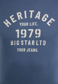 Zila kokvilnas T-krekls ar baltu, nodilušu tekstu, kurā ir "MANTOJUMS," "1979," un "BIG STAR LTD" izliekts līknes formā. Vienkāršs, ikdienišķs dizains.