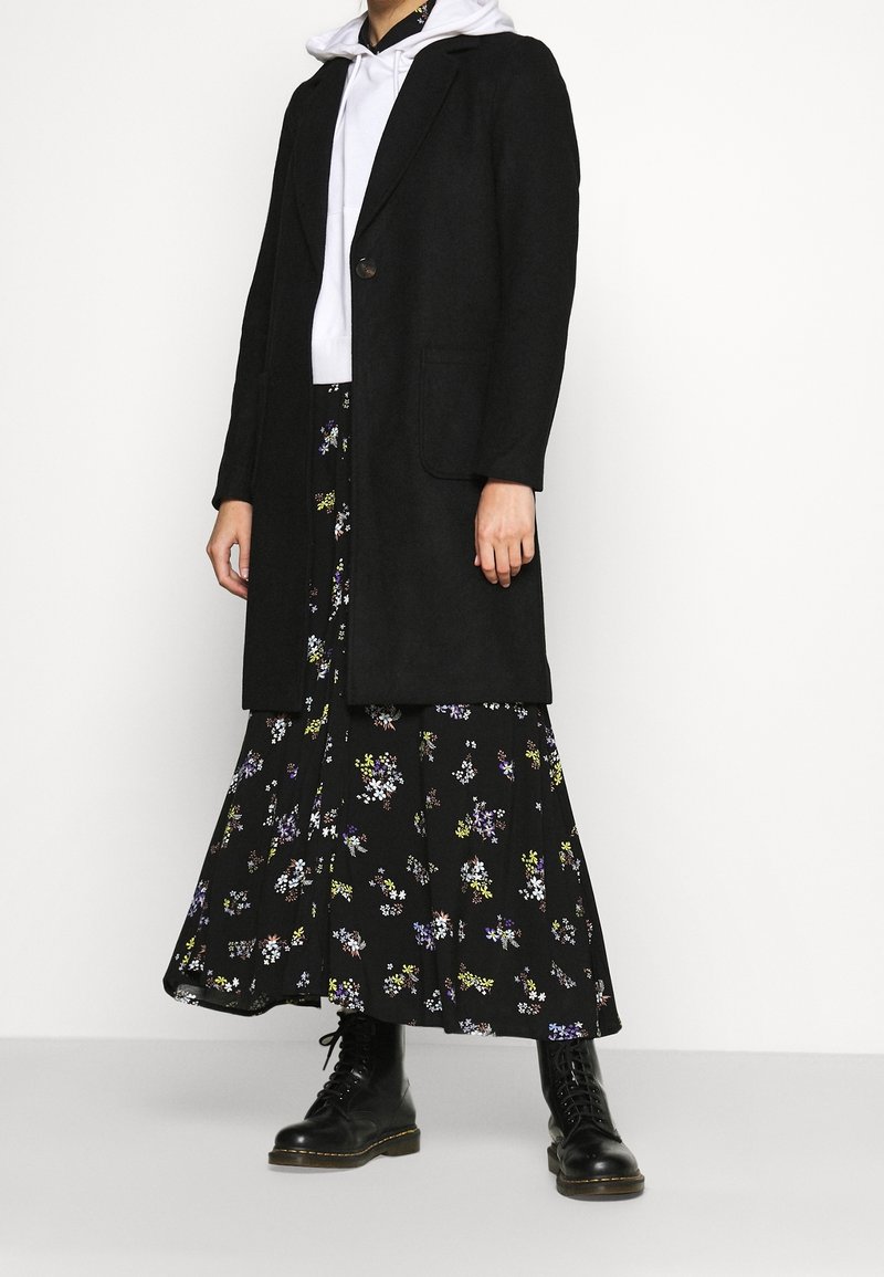 Personne portant un long manteau noir par-dessus un sweat à capuche blanc, une jupe longue noire fleurie et des bottes noires à lacets, debout devant un fond blanc.