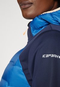 Colete acolchoado com frente azul e laterais azul-marinho. Apresenta um capuz ajustável, detalhes em zíper e o logótipo "ICEPEAK" em branco no ombro.
