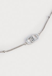 Bracciale in argento con un anello ovale che presenta un logo. La catena è delicata, con piccole perline e una superficie liscia.