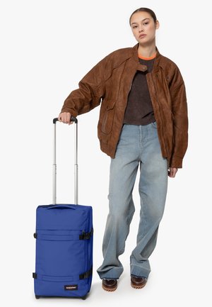Maleta azul con ruedas, dos cintas, tejido texturizado y un mango superior; sostenida por una persona que lleva una chaqueta de cuero marrón y jeans holgados.