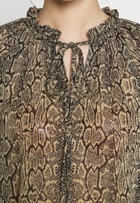 Gros plan d'une personne portant un chemisier beige et marron à imprimé serpent avec un col à volants et une encolure fendue nouée.