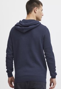 Sudadera color azul marino hecha de un material de punto texturizado, presenta una gran capucha, mangas largas y puños y dobladillo acanalados, vista desde la parte de atrás.