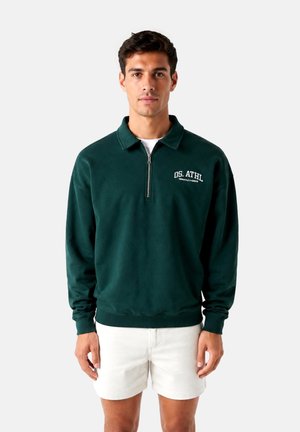 Mann, der einen dunkelgrünen Sweatshirt mit Viertel-Reißverschluss und "OS. ATHL" Text sowie weißen Shorts trägt, steht vor einem schlichten hellen Hintergrund.