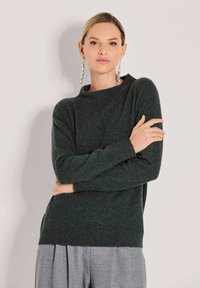 Frau mit blonden Haaren trägt einen dunkelgrünen Pullover, graue plissierte Hose und lange silberne Ohrringe, stehend vor einem schlichten hellen Hintergrund.