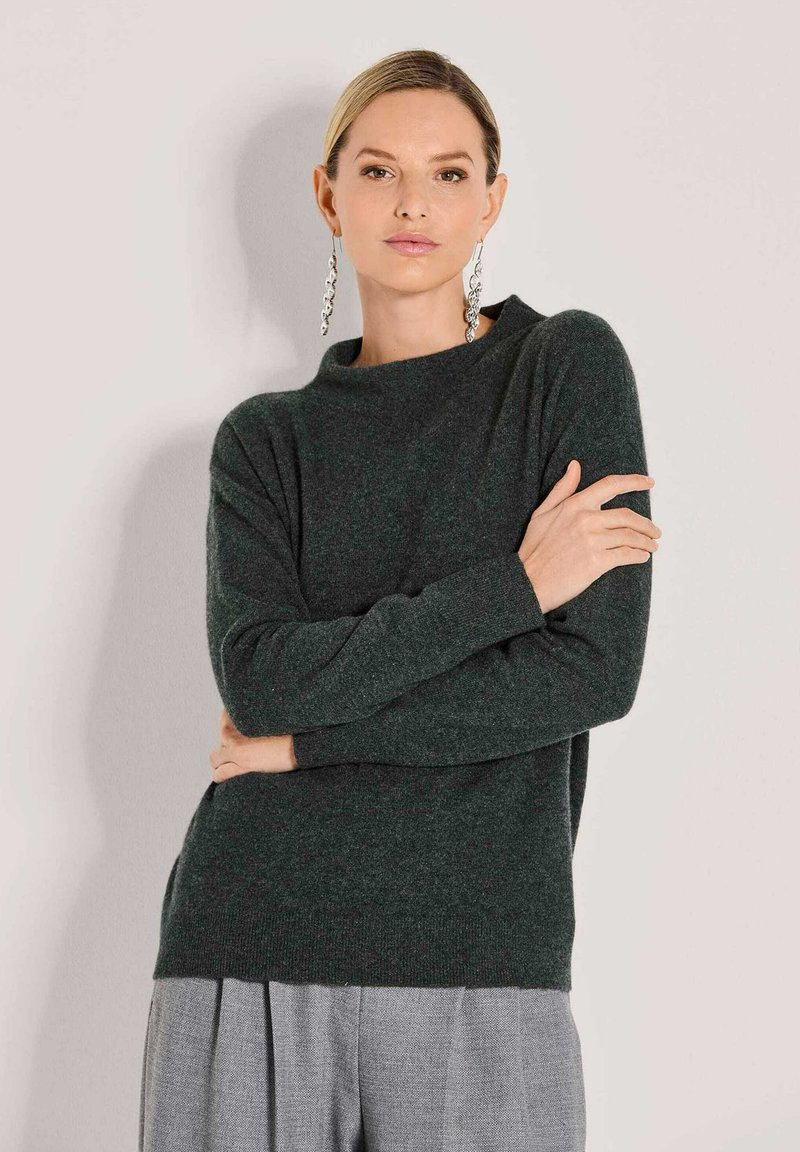 Frau mit blonden Haaren trägt einen dunkelgrünen Pullover, graue plissierte Hose und lange silberne Ohrringe, stehend vor einem schlichten hellen Hintergrund.