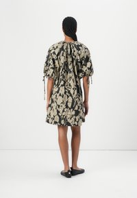 Femme aux cheveux tressés portant une robe noire avec un motif floral beige, des manches bouffantes avec des liens, et des chaussures noires sans lacets, debout de dos.