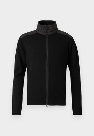 Melnā sporta jakā ar augstu apkakli, kurai ir strukturēti melni plecu paneļi un ribotas aproces un apakšmala. Visā jakā ir gluda auduma virsma.