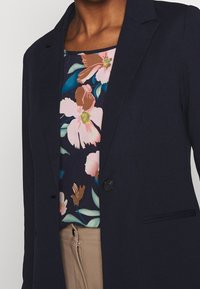 Blazer bleu marine à un bouton avec des revers crantés, porté sur un haut à imprimé floral avec des nuances de rose et de vert, associé à un pantalon beige.