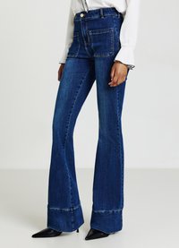 Hochgeschnittene, ausgestellte Jeans aus dunkelblauem Denim, mit zwei vorderen Taschen und einem Saum mit sichtbarer Naht, kombiniert mit schwarzen spitzen Absätzen.