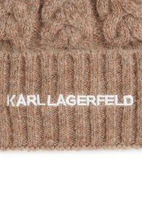 Hnedá pletená čiapka z textúrovanej priadze, s rebrovaným vzorom a výrazným bielym logom "KARL LAGERFELD" vpredu.