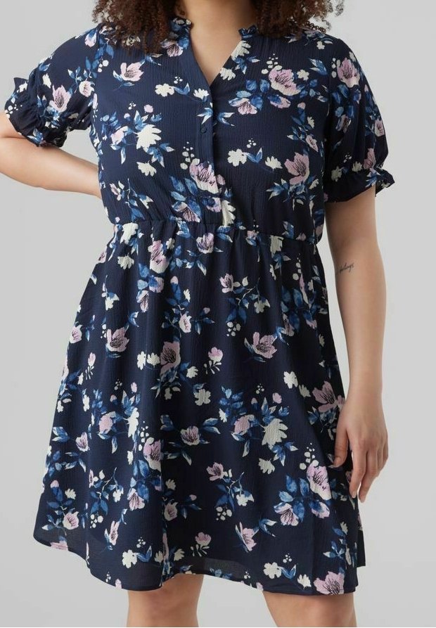 Femme portant une robe bleu marine à fleurs, manches courtes bouffantes et taille froncée, debout devant un fond gris uni.