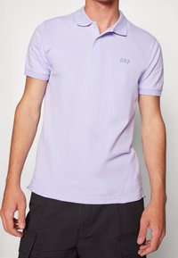 Polo de couleur violet clair en coton. Il est doté d'un col, de manches courtes et d'un petit logo "GAP" brodé sur la poitrine.