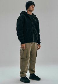 Czarny polarowy hoodie na zamek, beżowe spodnie cargo z bocznymi kieszeniami i czarne trampki. Model stoi na neutralnym tle.
