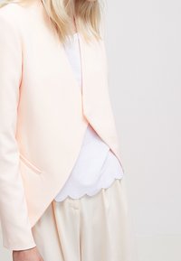 Blazer w kolorze brzoskwiniowym z opadającym, otwartym przodem, wykonany z gładkiego materiału i z minimalnymi detalami. Noszony na tle białej bluzki z falbanami u dołu oraz jasnych spodni.