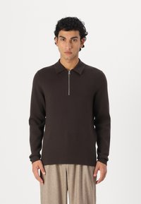 Brown knitted pullover with a zippered collar, long sleeves, and a straight hem worn with light-colored pleated trousers.