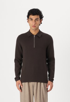 Brown knitted pullover with a zippered collar, long sleeves, and a straight hem worn with light-colored pleated trousers.
