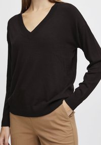 Schwarzer V-Ausschnitt-Pullover mit langen Ärmeln, gestrickter Textur und gerippten Bündchen, getragen über beigen Hosen. Einfaches Design ohne Muster oder Verzierungen.