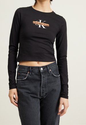 Femme portant un top court noir à manches longues avec le logo "Calvin Klein Jeans" et un jean gris foncé taille haute sur un fond uni.