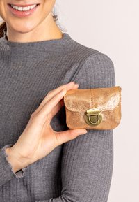 Kleine, gouden metallic leren pouch met een ronde vorm, gladde textuur en een ronde messing sluiting. Vastgehouden in een hand tegen een grijze trui.