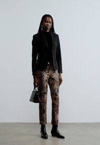 Blazer nero, top nero, pantaloni a fantasia con design floreale in beige e marrone. Borsa nera, stivaletti neri, sfondo neutro.