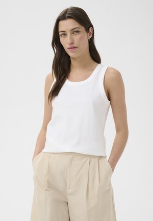 Femme aux longs cheveux bruns portant un haut blanc sans manches et un pantalon beige plissé, debout les mains dans les poches.