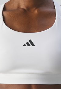 Vit sport-bh med rund halsringning, prydd med en svart Adidas-logotyp. Slät textur och minimalistisk design.