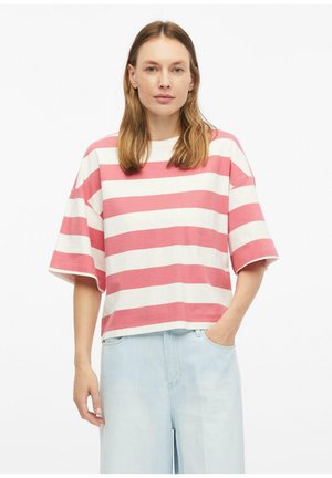 Kobieta o prostych, do ramion włosach, ubrana w przesadnie duży różowo-biały t-shirt w poziome paski oraz jasnoniebieskie szerokie spodnie jeansowe.