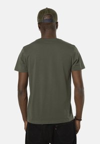 Hombre con una camisa de manga corta verde oliva y una gorra de camionero de malla verde, de espaldas, con pantalones negros y pulseras en la muñeca izquierda.