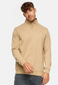 Pull beige avec un col montant et une fermeture à quart. Manches et ourlet côtelés, tissu doux avec une finition texturée, logo discret sur la poitrine.