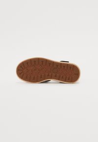 Chaussures noires avec une semelle en caoutchouc brun texturé. La semelle présente des motifs en flèche et en diamant, offrant adhérence et traction.