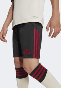 Niño que lleva pantalones cortos negros con rayas rojas, una camiseta blanca y calcetines altos blancos con bandas negras y rojas.