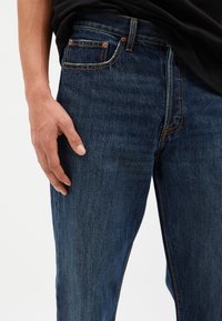 Vaqueros de mezclilla azul oscuro con corte recto, costuras claras a lo largo de las costuras y un bolsillo delantero con remaches metálicos.