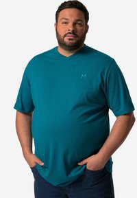 Tyrkysové tričko s výstřihem do V vyrobené z měkkého materiálu; má krátké rukávy a decentní logo na hrudi. Nosené s tmavě modrými džínami.