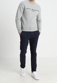Mann trägt einen grauen Tommy Hilfiger Pullover, marineblaue Hose und weiße Sneakers, steht mit den Händen in den Taschen vor einem weißen Hintergrund.