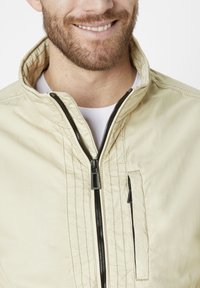 Light beige zip-up jacket with a high collar, black zipper, and a left chest pocket. Features visible stitching details and a smooth texture.