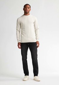 Pulover crem deschis cu decolteu rotund, purtat cu blugi negri. Modelul stă pe un fundal simplu, purtând adidași albi.