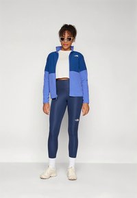 Niebieska kurtka z polaru z blokami kolorów, biały krótki top, granatowe legginsy i jasne sneakersy. Kurtka i legginsy ozdobione logo.