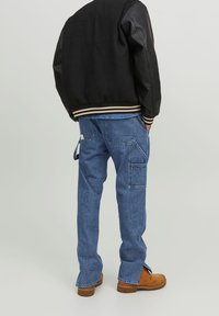 Chaqueta bomber negra con mangas de cuero, puños a rayas; pantalones cargo de mezclilla azul con bolsillos; botas marrones con cordones. Vista trasera.