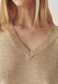 Beige gebreid sweater met een V-hals en een geribde rand. De stof lijkt zacht met een subtiele textuur en een lichte kleurvariatie.