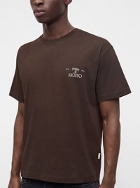 Homme portant un t-shirt marron uni à manches courtes avec le texte blanc "Treasure & Archivo" sur la poitrine gauche, sur un fond clair.